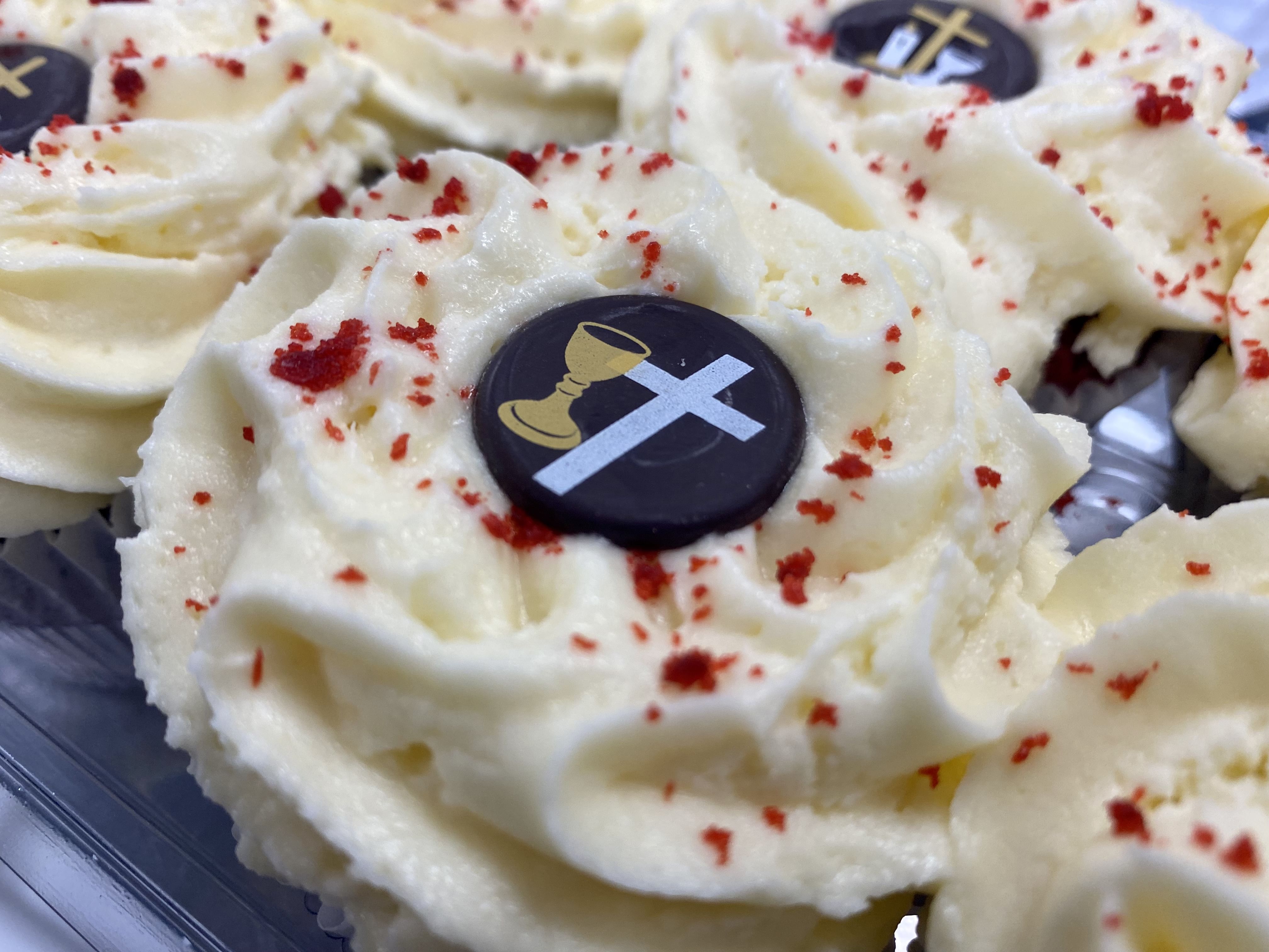 Close-up shot of Red Velvet Cupcakes with creamy white cream cheese frosting, sprinkled with red crumbs. Each cupcake is topped with a chocolate disc featuring a golden chalice and a white cross.